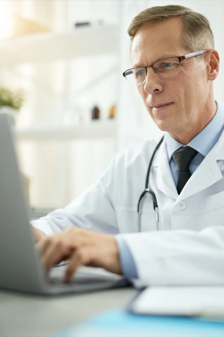 A physician reviewing records on a laptop at a desk